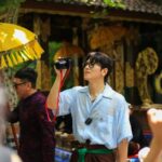 Ji Chang Wook holding a camera, exploring a cultural site in Bali, Indonesia, surrounded by traditional decorations and umbrellas.